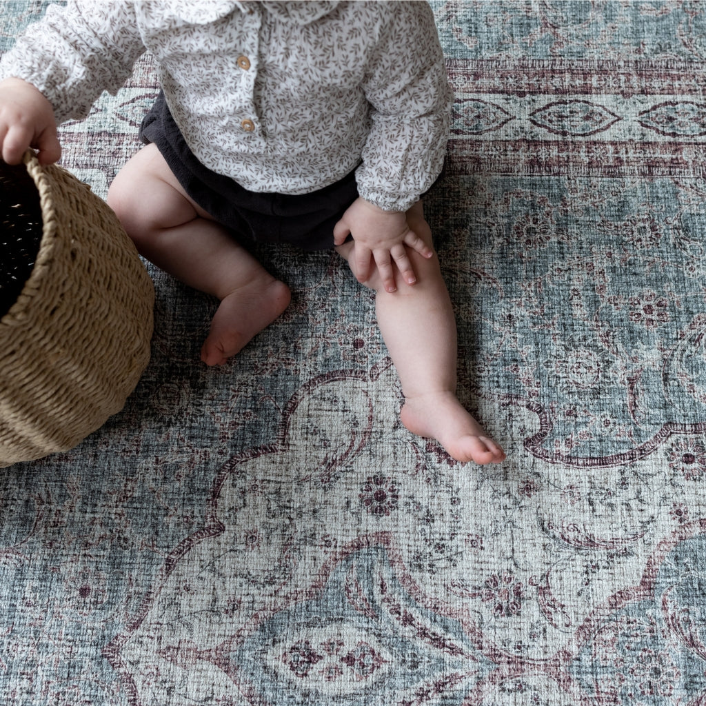 A baby sits on a memory foam play mat rug in deep red and blue-gray tones.