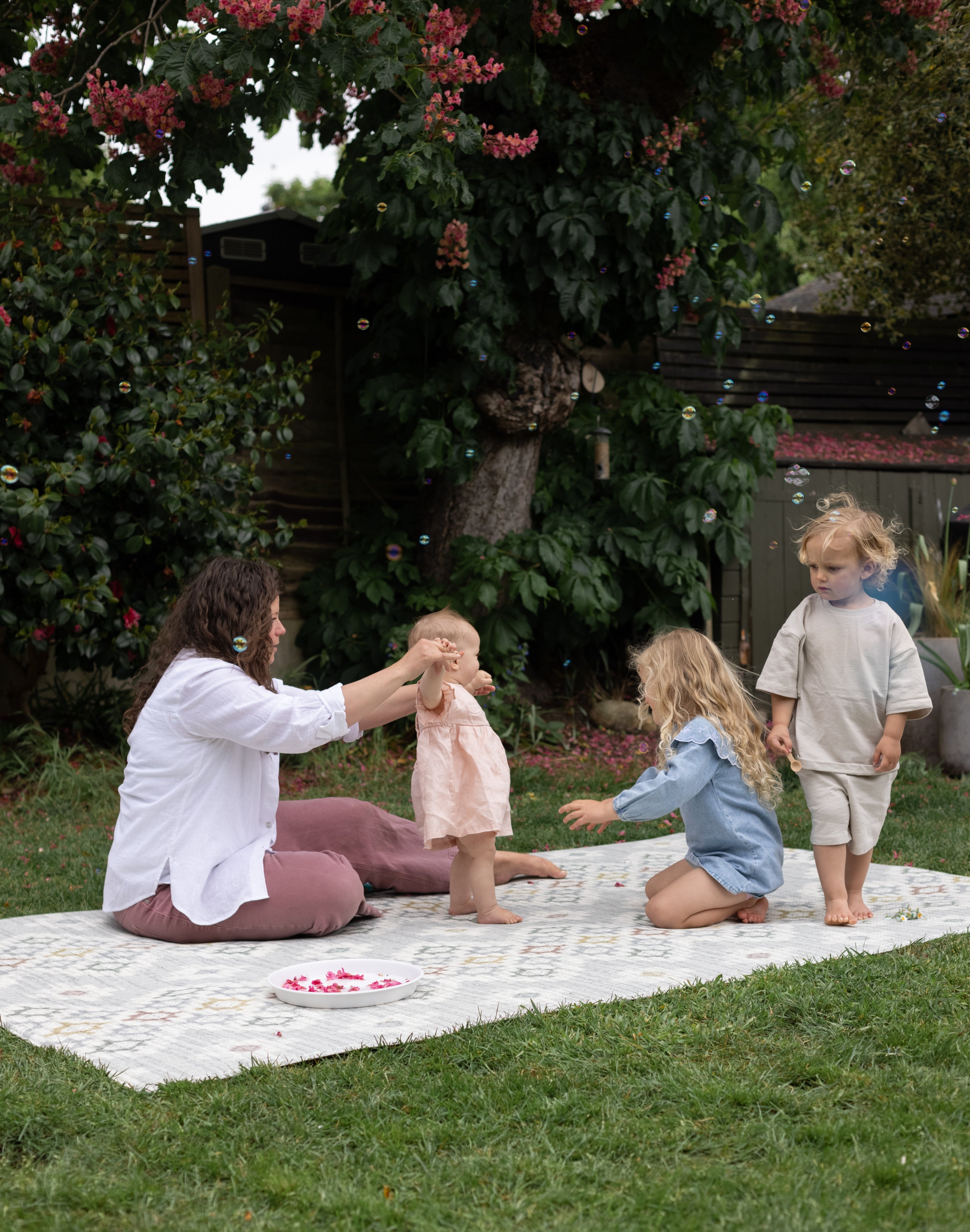 Woman and children playing outdoors on a wipeable memory foam play mat with trees and greenery in the background