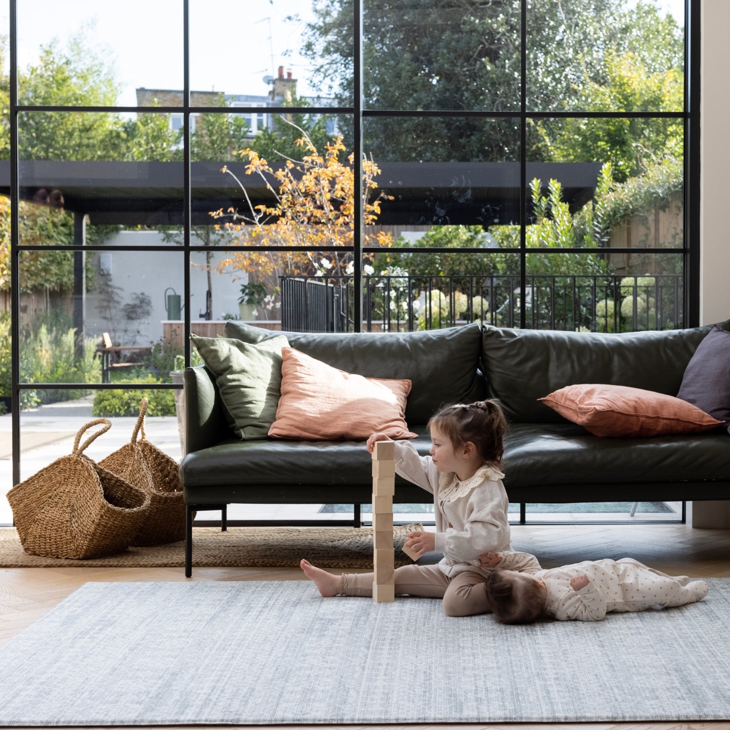 A family room scene. A toddler plays with stacking blocks while a baby lies down looking comfortable. They are on a large grey patterned play mat for infants