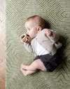 Little baby chews on wooden teether while being supported on a cushy floor mat designed for the whole family