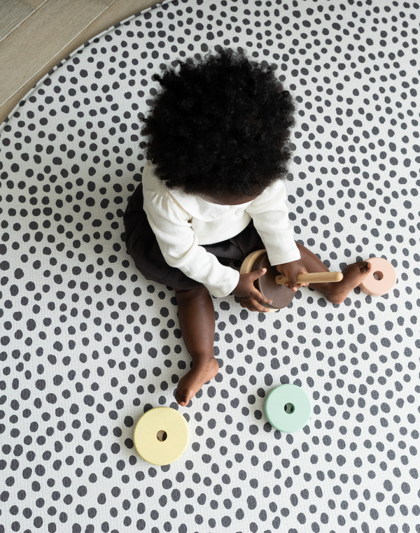 Baby girl enjoys playtime with a wooden stacking toy protected on the floor with the round foam play mat
