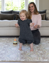 Adorable toddler with limb difference standing on the supportive memory foam play mat by Totter and Tumble with her mother sitting behind smiling as she watches 