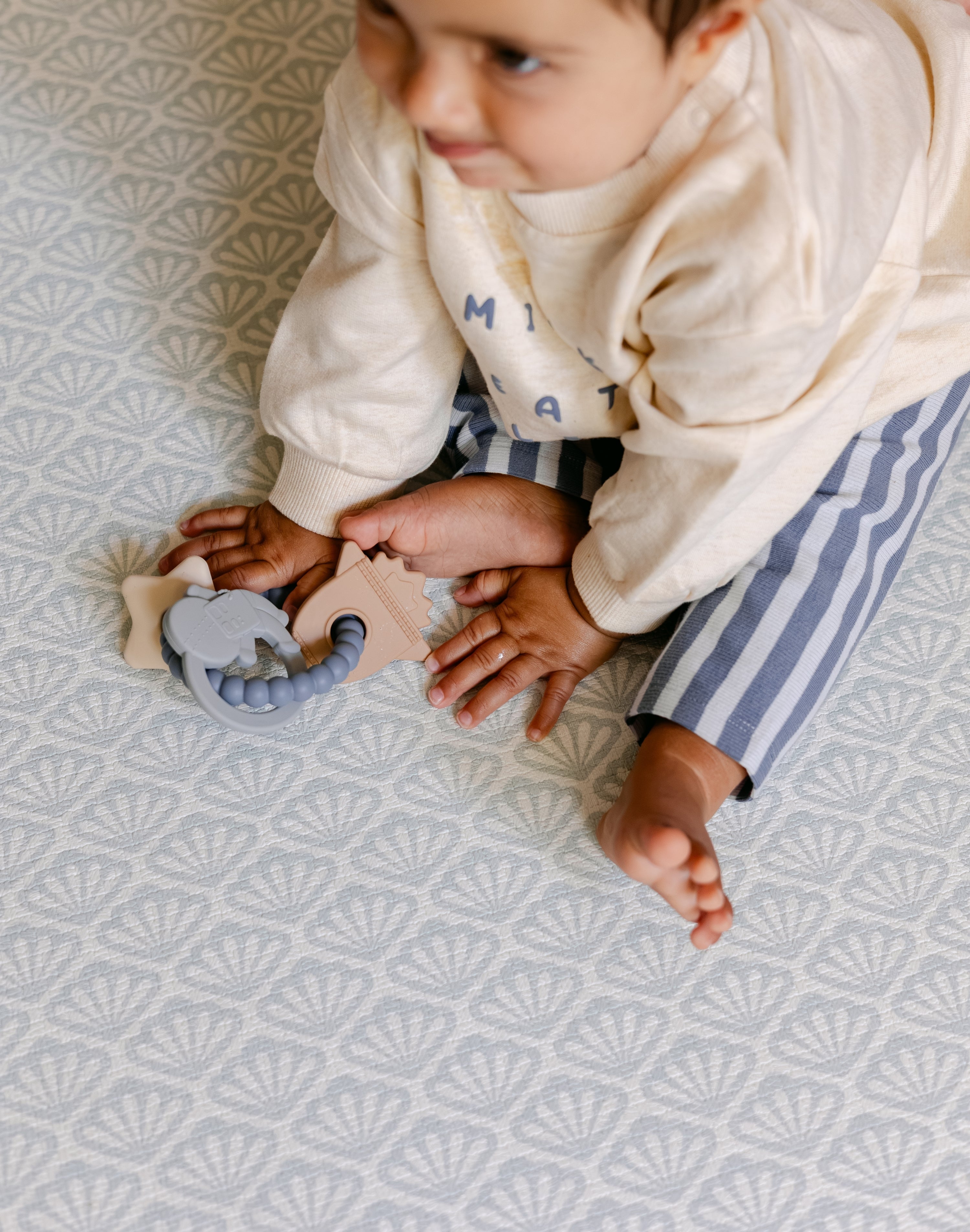 A baby sat comfortably on The Beachcomber play mat. With a repeating shell design. | SKU: 26 BEACH/GATHER XL, 26 BEACH/GATHER STD, 26 BEACH/GATHER COMP