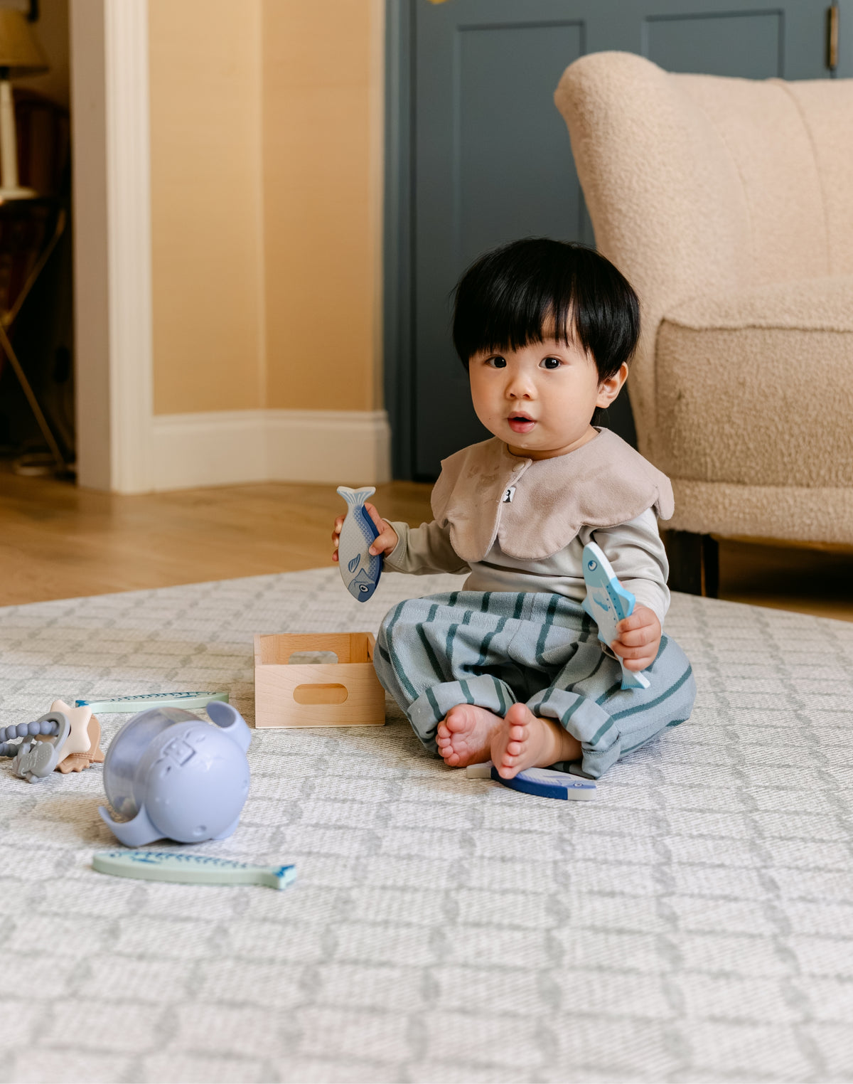 A toddler sits barefoot on a patterned memory foam totter and tumble play mat in a living room, holding two light blue toy fish. The child is wearing a beige bib and green striped pants.