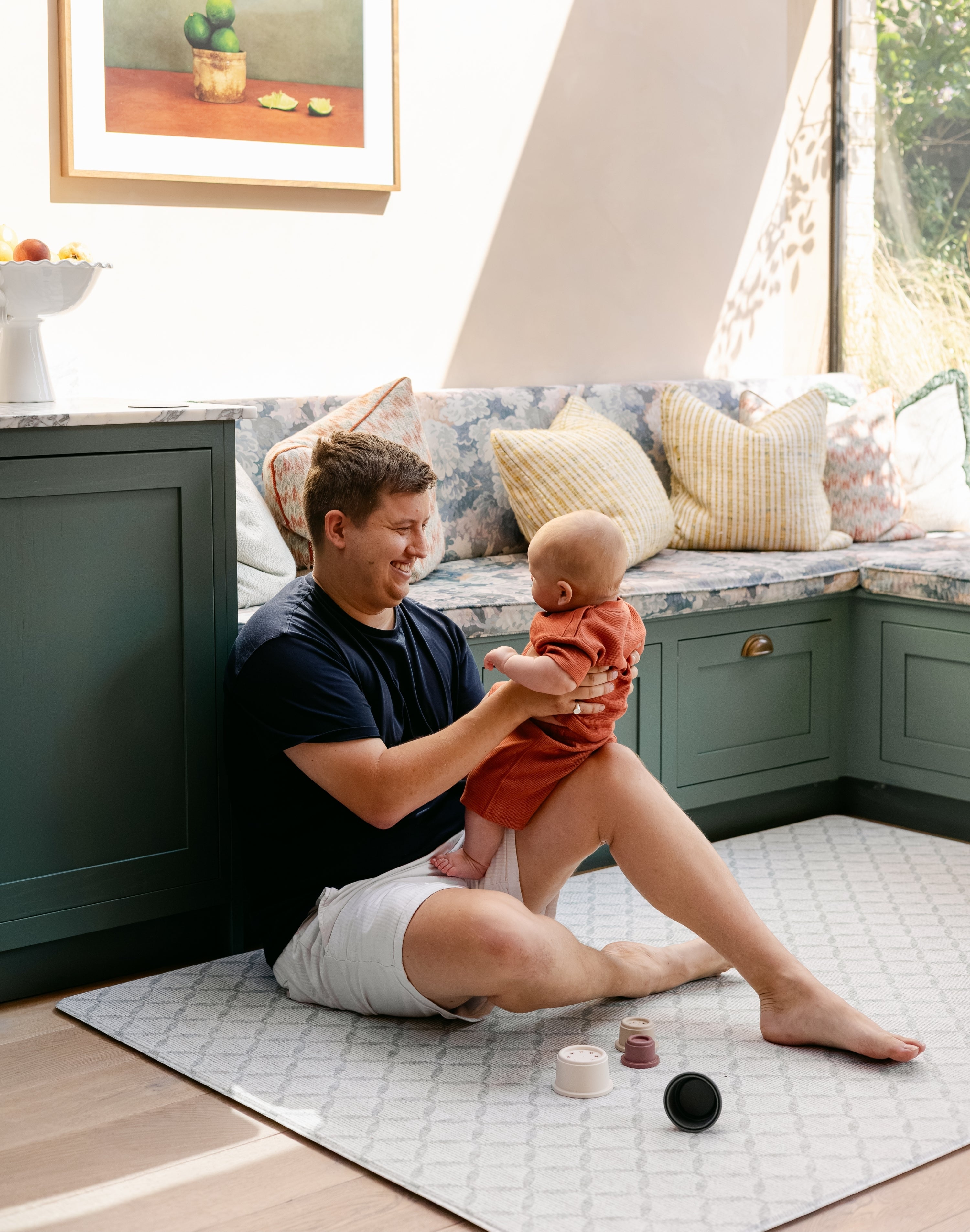 A man sat comfortably holding a baby on the gatherer play mat, made from thick safe non toxic memory foam | SKU: 26 BEACH/GATHER XL, 26 BEACH/GATHER STD, 26 BEACH/GATHER COMP